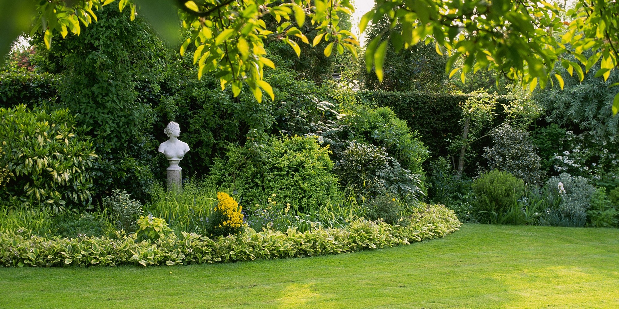 Een deel van een tuin met gazon, een border bestaand uit struiken en bomen en een beeldje