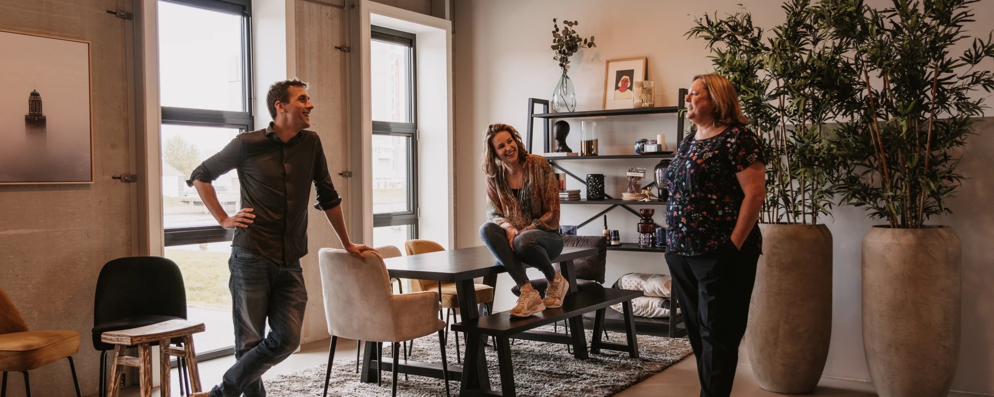 Informeel gesprek tussen drie mensen in een moderne, ontspannen kantooromgeving met een tafel, decoratieve elementen en kamerplanten op de achtergrond.