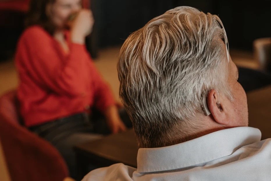 Man met grijs haar en hoortoestel zit met rug naar de camera; vrouw op de achtergrond in gesprek, onscherp in beeld.